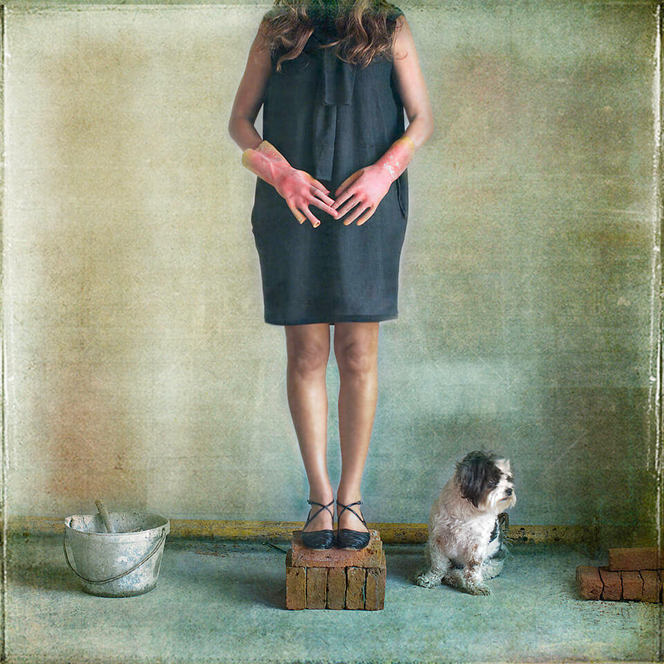 Eine Frau steht auf gestapelten Backsteinen, ein kleiner Hund sitzt daneben.