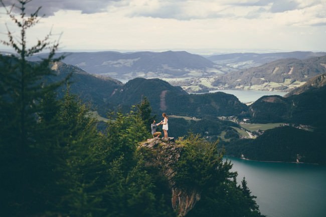 Ein Mann kniet vor einer Frau, sie stehen auf einem Vorsprung in einer weiten Seen- und Gebirgslandschaft.