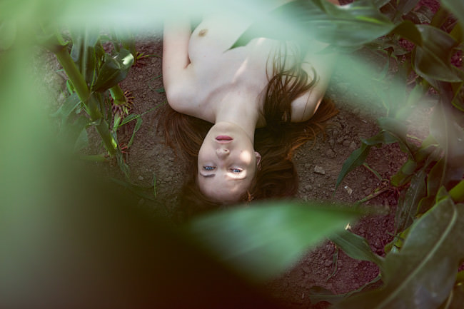 Oberkörper einer nackten Frau auf dem Boden liegend, durch grüne Blätter fotografiert.