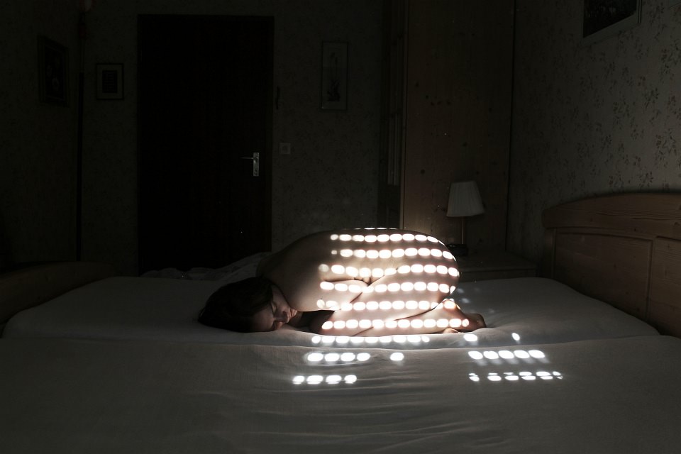 Nackte Frau zusammengekauert auf einem Bett, in einem dunklen Zimmer, auf ihrer Haut Lichtflecken, die durch einen Rollladen fallen.
