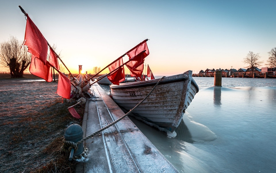 Am Steg festgefrorenes Boot mit roten Fahnen im morgendlichen Gegenlicht.