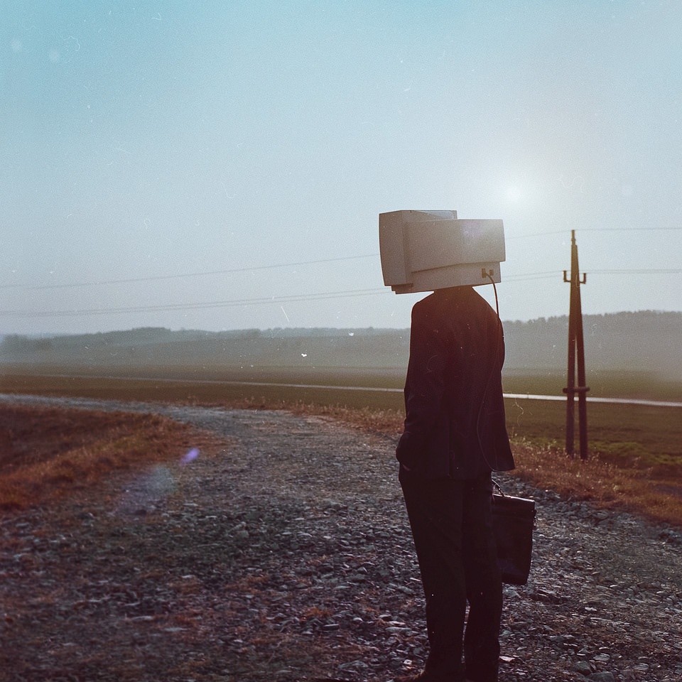 Mann im Anzug mit Aktenkoffer und Computerbildschirm statt Kopf steht auf einer ländlichen Straße.