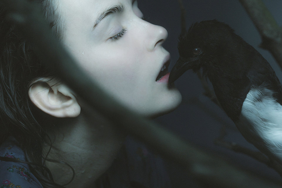 Ein bleiches Mädchen lässt sich von einem dunklen Vogelschnabel berühren.