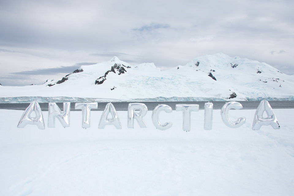 Silberne Luftballons mit den Buchstaben Antarctica vor Eisbergen.