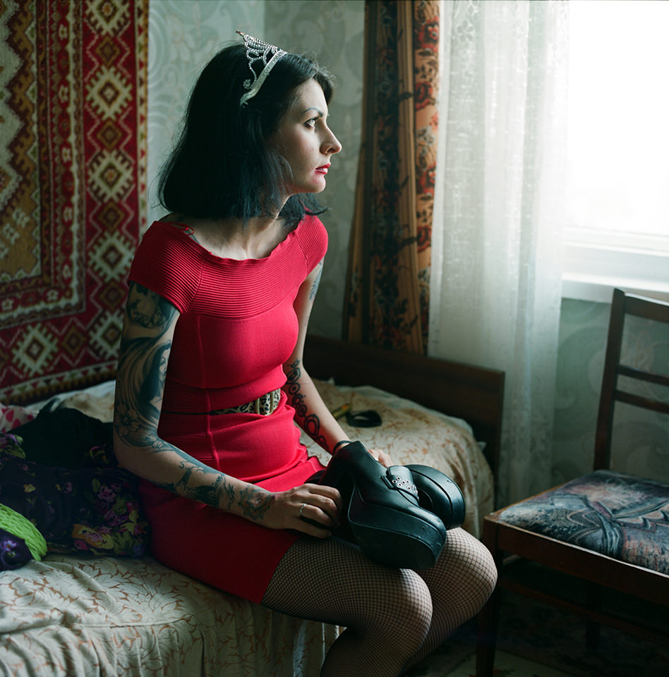 Eine tätowierte Frau in einem roten Kleid mit einer Krone und verschmierten Lippenstift sitzt auf dem Bett und schaut aus dem Fenster