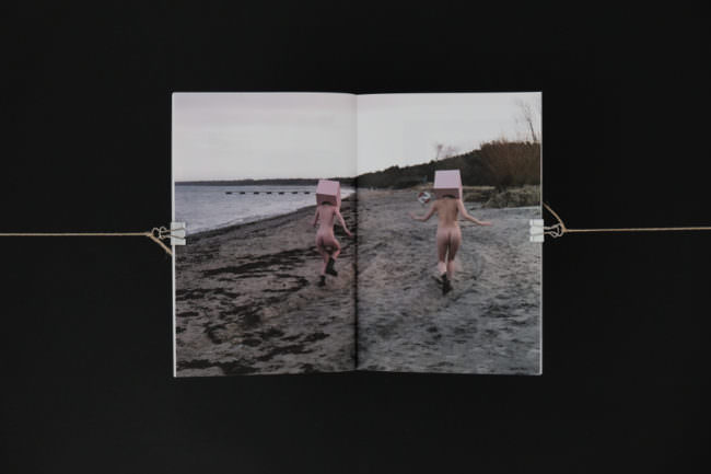Zwei nackte Frauen laufen mit Kartonköpfen über einen Strand.