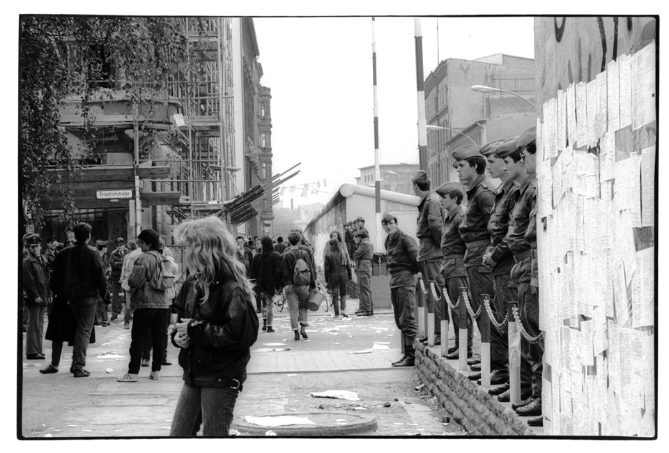Eine Frau steht vor einre Mauer und Grenzsoldaten.
