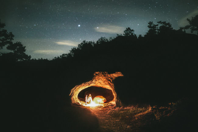 Zwei Personen stehen neben einem Lagerfeuer in einer Höhle unter Sternenhimmel