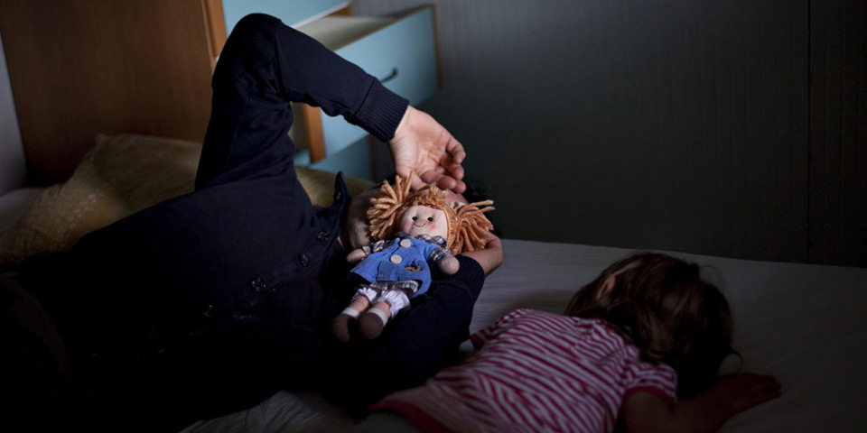 Ein Erwachsener und ein Kind liegen auf einem Bett, der Erwachsene hält sich eine rothaarige Puppe vors Gesicht.