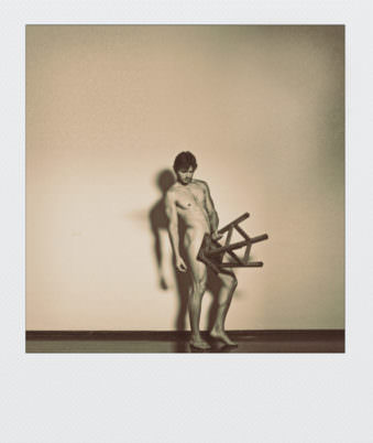 Polaroid eines nackten Mannes der seine Scham mit einem Stuhl bedeckt.