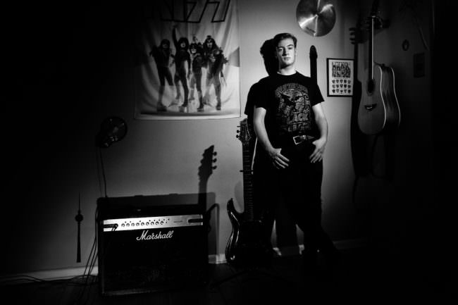 Junger Mann stehend neben Amp und Gitarre lehnt an Wand mit Bandposter.