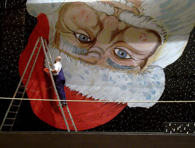Wandbild vom Weihnachtsmann auf dem Kopf, davon ein Mann auf einer Leiter in Blaumann.