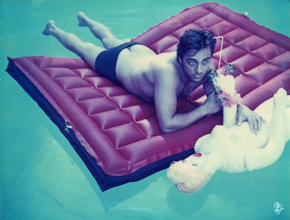Ein Mann liegt in einem Pool auf einer Luftmatraze,in seinen Händen ein Cocktail und eine Gummipuppe