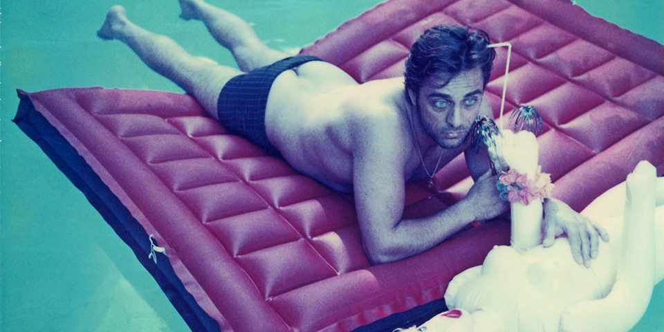 Ein Mann mit Gummipuppe und Cocktail liegt auf einer Luftmatraze im Pool.
