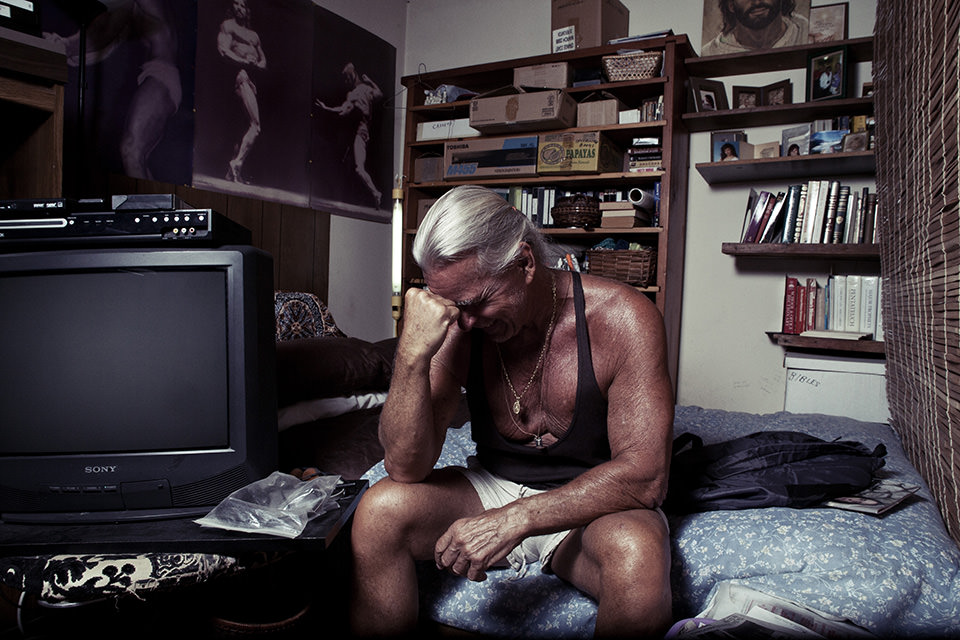 Mann im Muskelshirt und mit ergrauten langen Haaren sitzt auf einem Bett, scheinbar Niedergeschlagen.