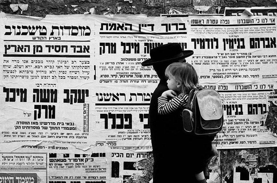 Eine Person trägt ein Kind an einer mit hebräischen Plakaten behangenen Wand vorbei