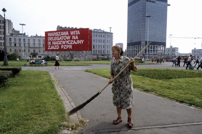 Eine ältere Dame mit Schürzenkleid fegt mit einem Reisigbesen einen Weg. Im Hintergrund sind städtische Gebäude zu sehen.