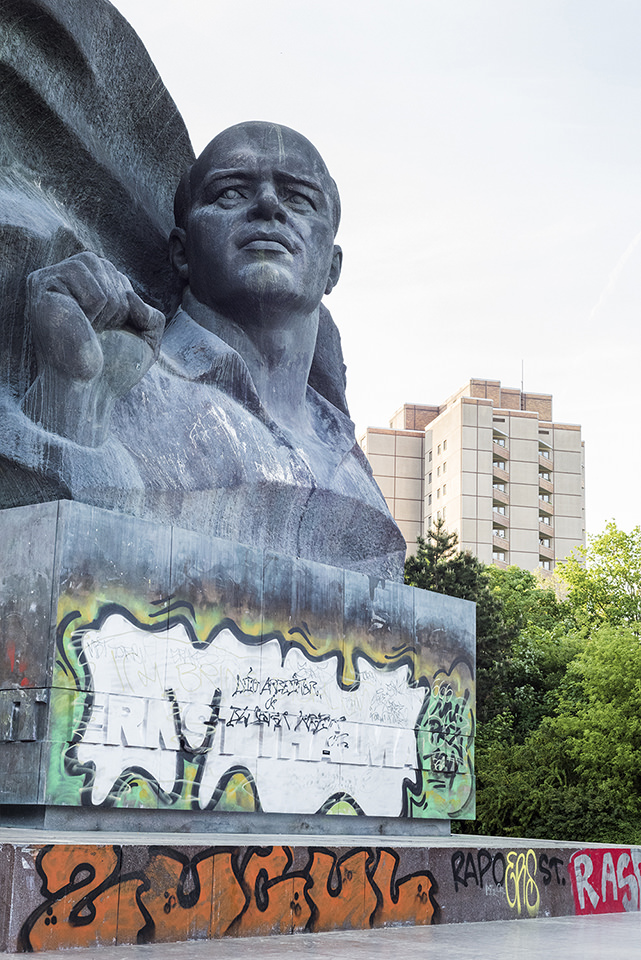 Denkmal Staue im Vordergrund mit Graffiti bemalt, dahinter ein Hochhaus und Grünwuchs.