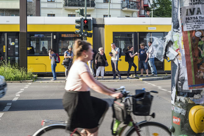 Frau auf einem Fahrrad fährt vorn durchs Bild, im Hintergrund Personen die an einer Ampel warten. Hinten ihnen fährt eine Straßenbahn entlang.