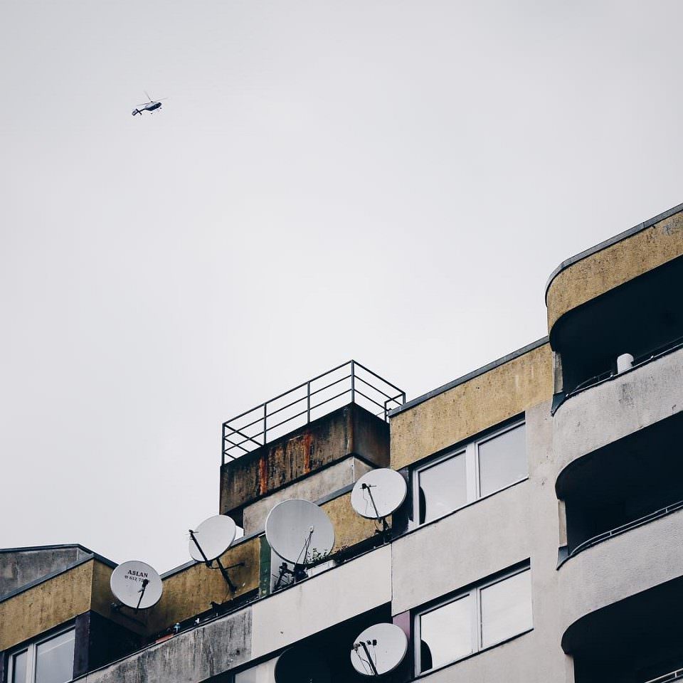 Fassade mit Balkonen, Fenstern und Satellitenschüsseln vor einem grauen Himmel, an dem ein Helikopter schwebt.