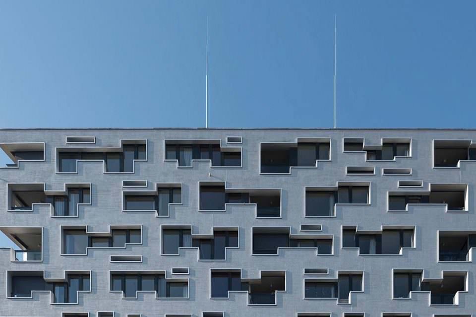 Moderne Fassade mit verwinkelten Fensteröffnungen, auf dem Gebäude zwei lange Antennen, vor einem blauen Himmel.