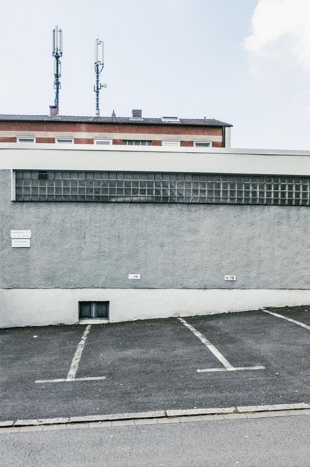 Schräg abfallender Parkplatz an der Rückwand eines grauen Gebäudes.