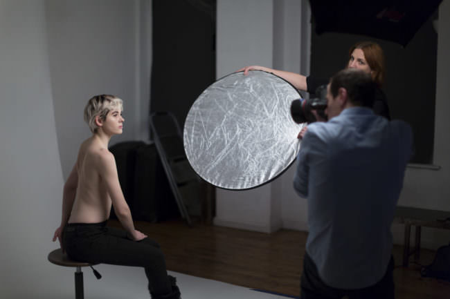Fotograf fotografiert sitzendes Modell, eine Assistentin hält einen Reflektor.
