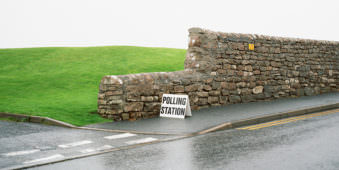 Natursteinmauer zwischen Wiese und Straße mit einem Schild auf dem Polling Station steht.