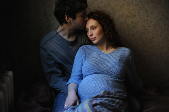 Junges Paar, der Mann küsst seine schwangere Frau auf die Stirn. Sie sitzen nebeneinander an eine Wand gelehnt. Die Frau hat einen blauen Pulli an.