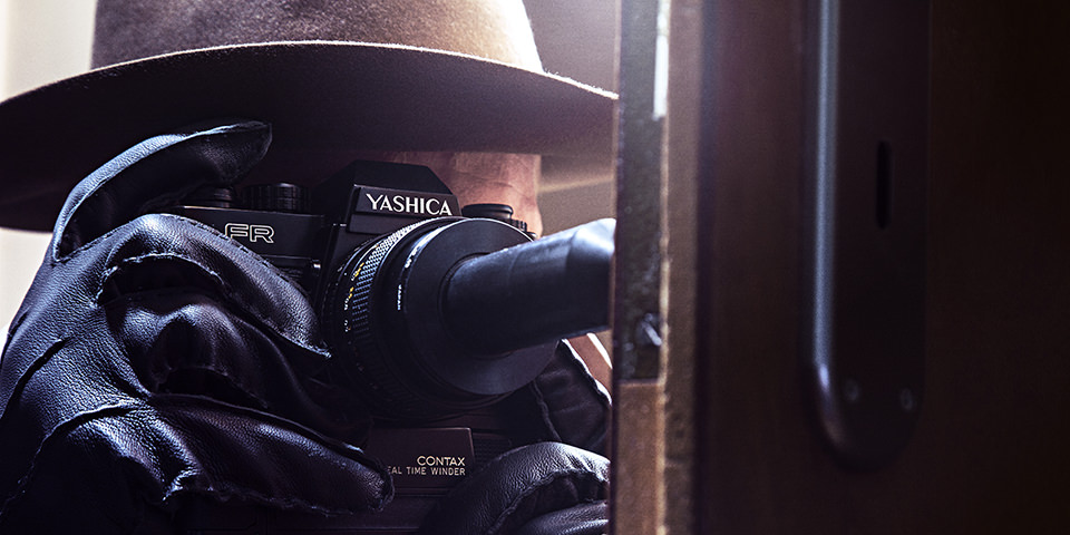 Spionagefotograf mit Hut und Lederhandschuhen fotografiert mit besonderer Kamera durch etwas hindurch.