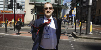 Mann mit Anzug der auf einen zuläuft, seine Jacke ist offen und die rote Krawatte weht im Wind. Er befindet sich in einer städtischen Umgebung.