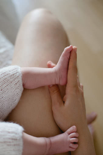 Ein Babyfuß am Zeigefinger einer Erwachsenenhand. Beide liegen auf einem nackten Erwachsenenbein.