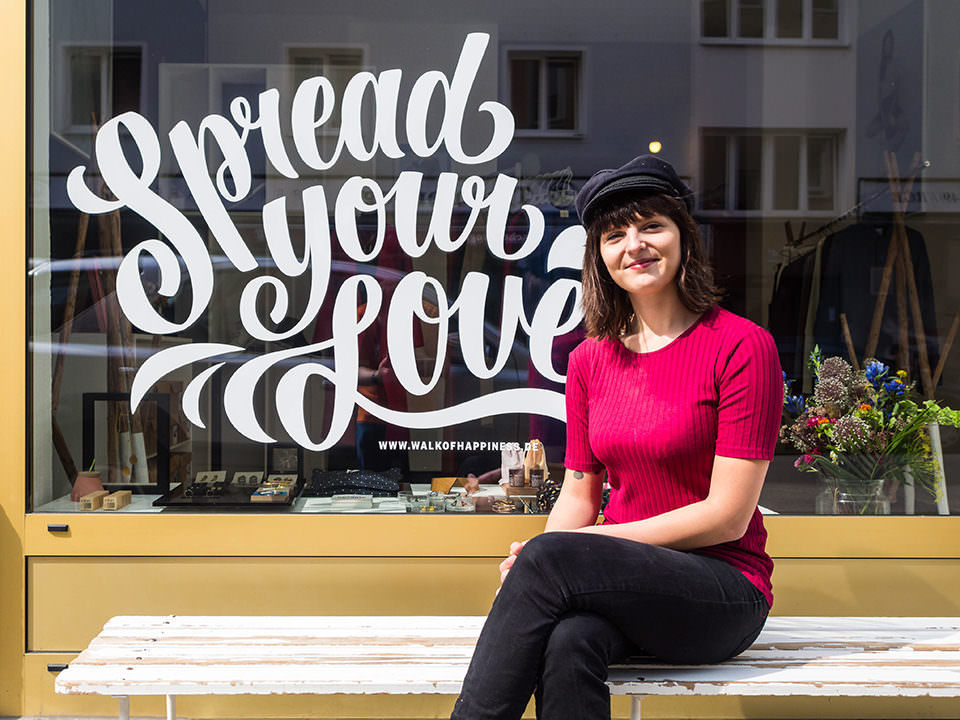 Junge Frau mit dunklen Haaren und einer Mütze auf sitzt vor einem Schaufenster auf dem in geschwungenen Buchstaben "Spread your Love" steht.