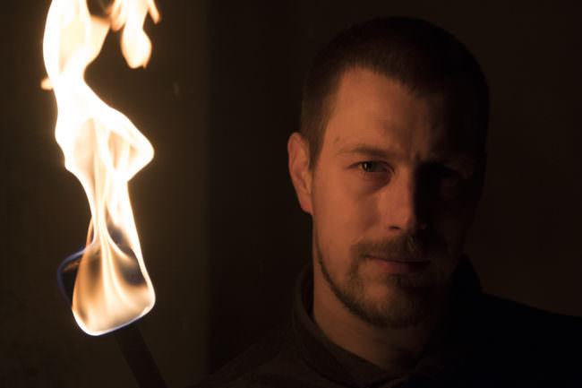 Portrait eines jungen Mannes, der nur von einer Flamme neben ihm beleuchtet wird.