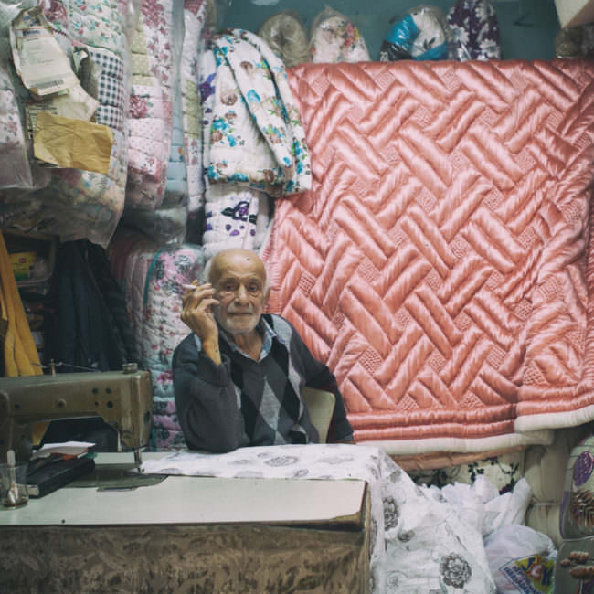 Rauchender Mann sitzt hinter einer Nähmaschine in einem kleinen Laden für Bettdecken.