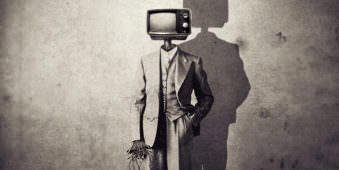 Person in Anzug mit Maschinenkopf als TVaparat mit Schatten vor einer gemusterten Wand.