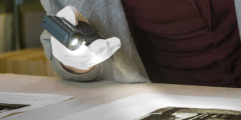 Hand in weißem Handschuh hält eine besonders geformte Lampe und leuchtet auf ein Bild.
