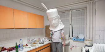 Person mit Pappmachée Kopf aufgesetzt in einer Küche.