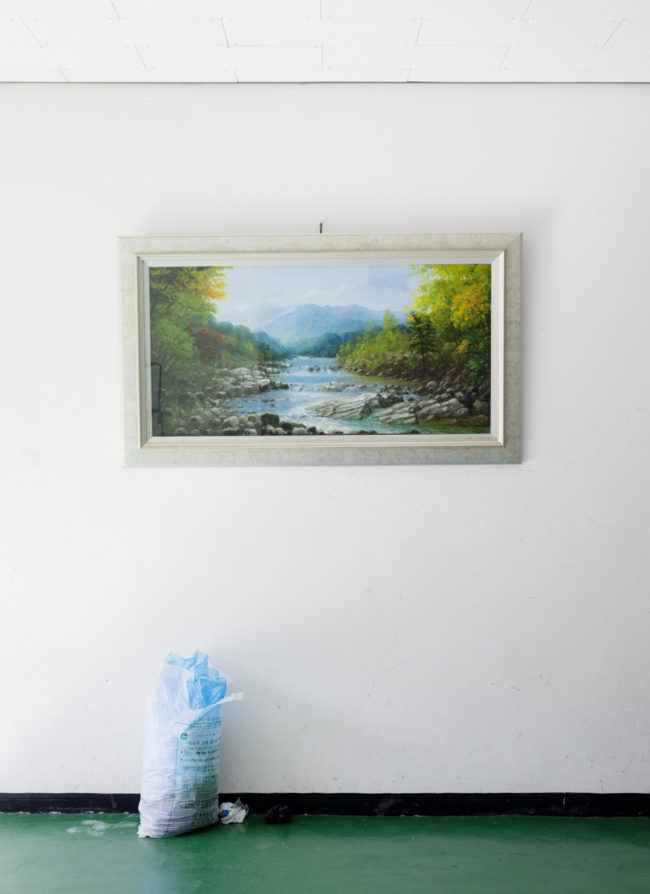 Ein gerahmtes Bild von einem Fluss hängt an einer Wand, darunter ein Müllsack.