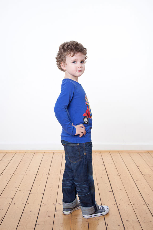 Ein Kind mit blauem Shirt stehend auf Holzfußboden vor weißer Wand.