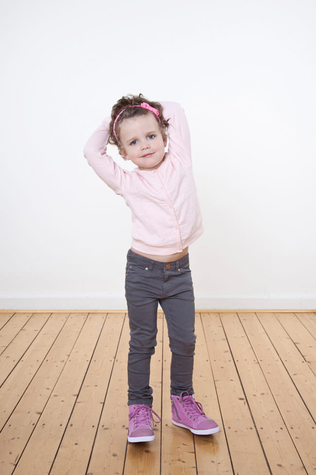 Ein Kind mit rosa Shirt stehend auf einem Holzfußboden vor einer weißen Wand.