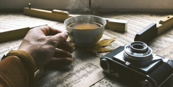 Eine Hand hält eine Tasse heißen Kaffee, die auf einem Schreibtisch voller Notenblätter steht. Daneben liegt eine Kamera.