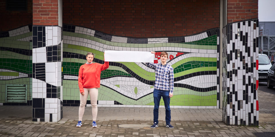 zwei Personen halten einen Banner vor einer bunten Wand hoch
