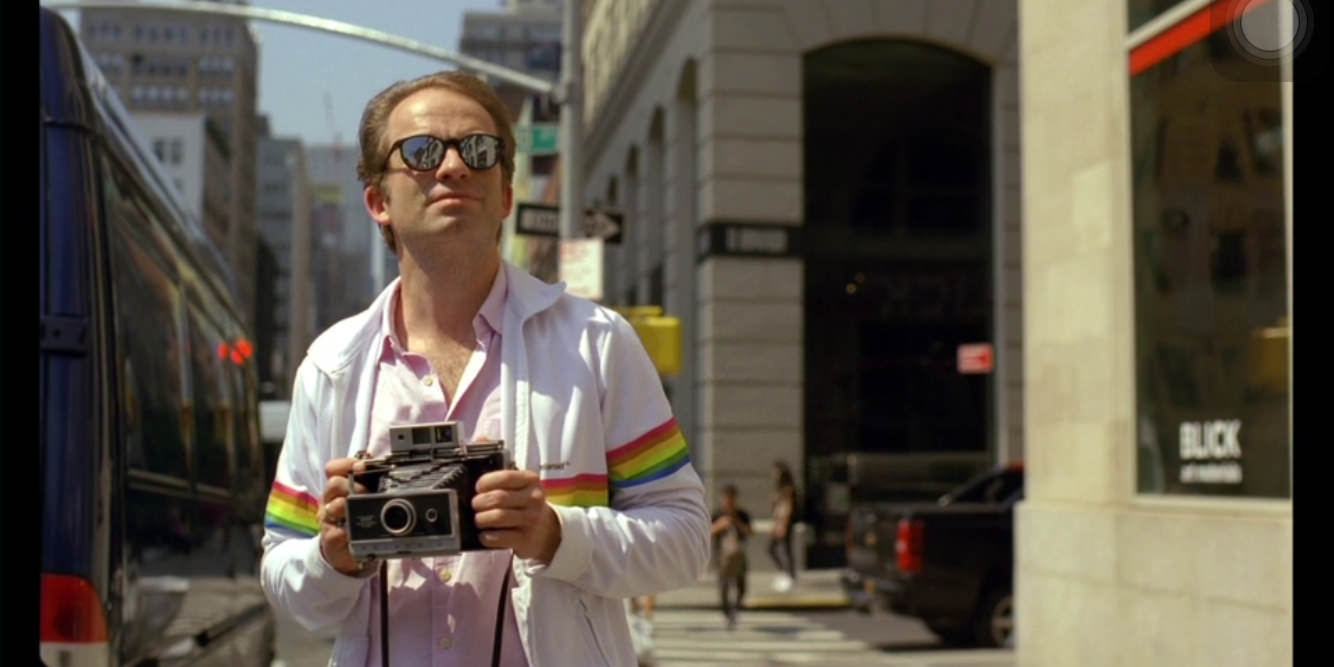 Filmscreenshot. Mann mit Polaroidkamera auf der Straße