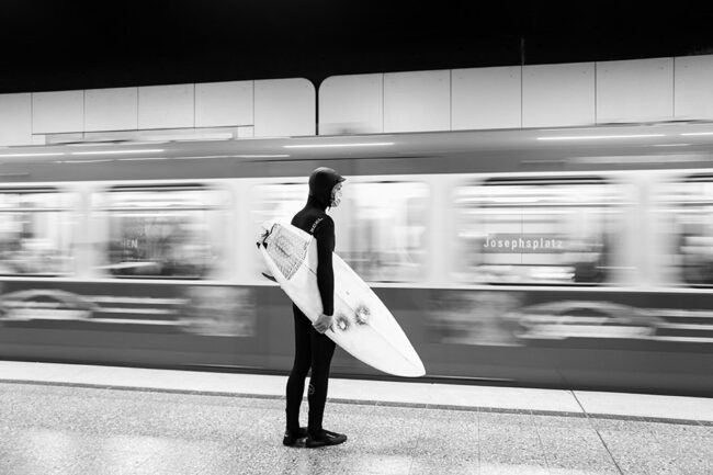 Ein Surfer steht mit Surfbrett und im Anzug an einer U-Bahnhaltestelle