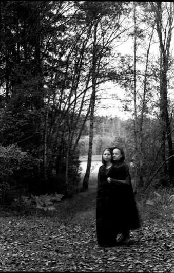 zwei sich umarmende Personen in Mänteln vor Bäumen an einem See