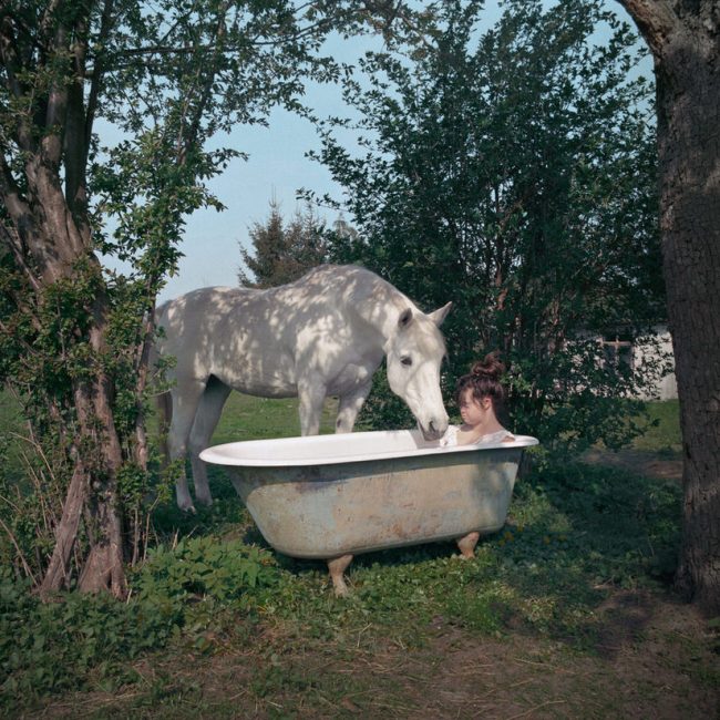 Frau badet draußen. Ein weißes Pferd steht daneben.