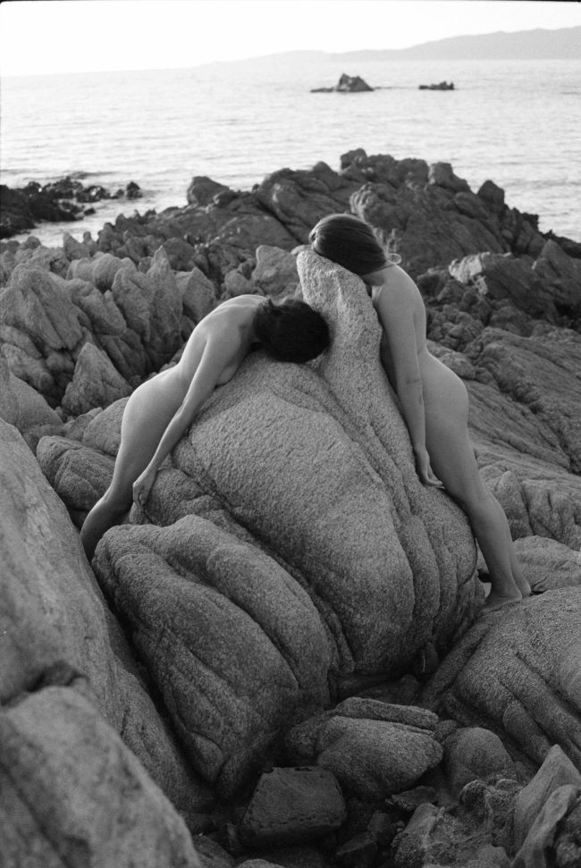 Zwei Personen lehnen nackt an einem Stein am Strand