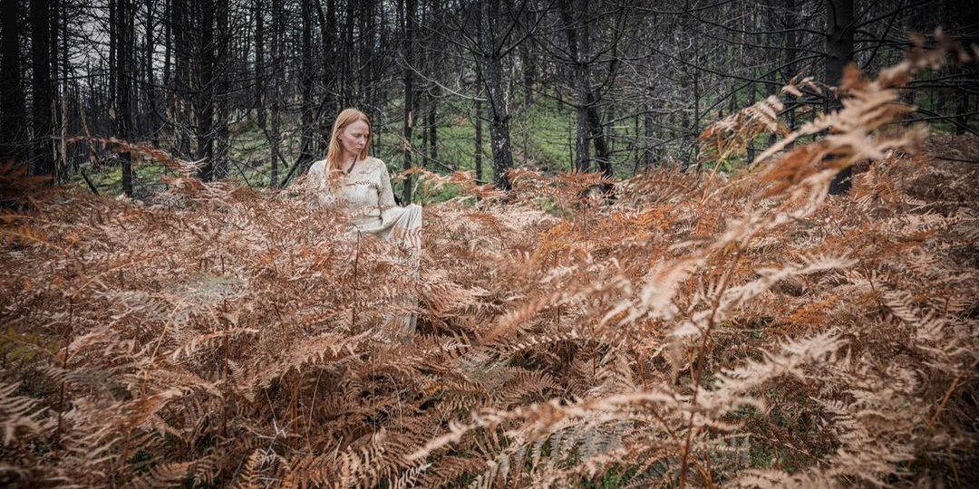 Frau in langem, hellem Kleid inmitten von herbstbraunen Farnen in einem Wald.