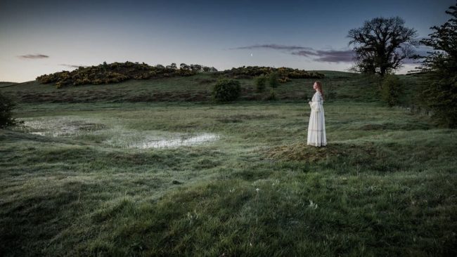 Frau in langem, hellem Kleid auf einer Wiese mit Sumpf und Hügeln im Hintergrund.